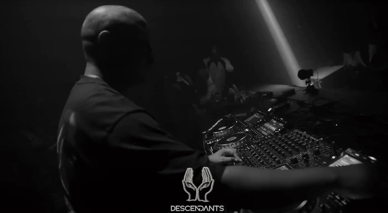 Un GIF en blanco y negro muestra al DJ Caiiro de perfil, concentrado en la mesa de mezclas durante un set en vivo. Sus manos se mueven sobre los controladores bajo una luz de foco. El logo del evento, "DESCENDANTS", es visible en la parte inferior.